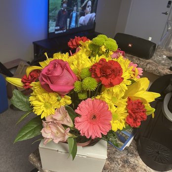 Crismon's Flowers - Flowers - A bouquet of mixed flowers, including pink roses, yellow lilies, red carnations, and pink gerbera daisies, is arranged in a container on a kitchen countertop. The flowers are fresh and colorful. In the background, a television displays a scene with people outdoors, and a black speaker is positioned on the counter. The setting appears to be an indoor kitchen or living area with various household items visible. - Florists Near Me - Mesa, Arizona