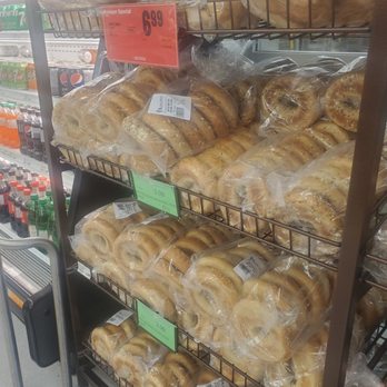 Adonis - Multiple bags of bagels are stacked on metal shelves in a grocery store. The bagels are in clear plastic packaging, and price tags are attached to the shelves. The background shows refrigerated beverages, including soda bottles and cans, indicating a typical grocery store setting. - Grocery Near Me - Toronto, Ontario