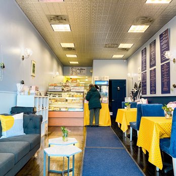 Bakehaus - Inside - A bakery interior features a seating area with blue chairs and yellow tablecloths, a display case with baked goods, and a customer at the counter. - Desserts Near Me - Belmont, Massachusetts