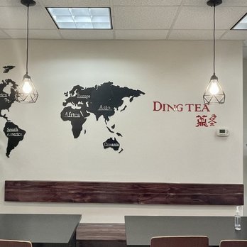 Ding Tea Rolling Meadows - The image shows the interior of Ding Tea, with a wall decorated with a black world map decal labeled with continents. Two pendant lights hang from the ceiling, illuminating the space. A potted plant is positioned on the left side, and a digital ordering kiosk is on the right. The seating area includes a table with chairs, and the wall displays the Ding Tea logo along with some promotional posters. - Bubble Tea Near Me - Rolling Meadows, Illinois