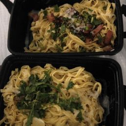 Lemon pasta and Carbonara Alexandra - Two black takeout containers hold pasta dishes, one topped with chopped green herbs and the other with bacon bits and green onions.