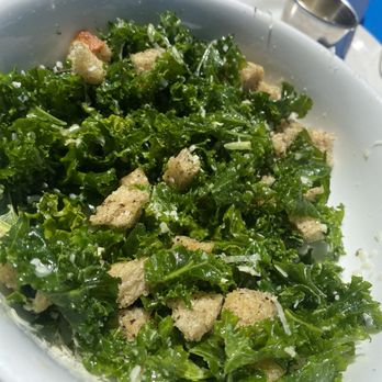Le Paddock - $15 Kale "Ceaser" except there's no dressing and the croutons were stale and tasteless. - A fresh kale Caesar salad with croutons is shown in a white bowl placed on an outdoor table. The salad contains chopped kale, croutons, and grated cheese, with sunlight illuminating the dish. In the background, there is a blue surface and a metal cup, indicating an outdoor dining setting. - French Near Me - Brooklyn, New York