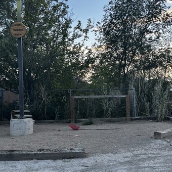 Village Pizza - "Horse parking"area - The image shows a gravel parking area with a tall lamp post on the left side, which has a circular sign attached that reads 'Horse Parking Only!!!'. The parking lot is bordered by a wooden fence and dense trees and bushes, with a partly cloudy sky overhead. The ground appears uneven with some wooden planks and a red object lying on the gravel. - Pizza Near Me - Corrales, New Mexico