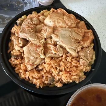 Phoenix Coqui - Chicken Mofongo Bowl - A black bowl contains rice topped with cooked chicken pieces and vegetables. Surrounding it are containers of sauce and a bowl of soup, on a light-colored table. - Puerto Rican Near Me - Phoenix, Arizona