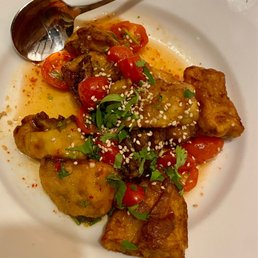 Crispy Eggplant - A food plate contains crispy fried eggplant pieces garnished with cherry tomatoes, chopped herbs, and sprinkled sesame seeds. The dish appears to be part of a Central Asian cuisine, served on a white ceramic plate with a metal spoon nearby. The presentation emphasizes fresh ingredients and vibrant colors.