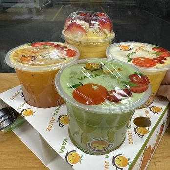 Vara juice - Four fruit smoothies in clear plastic cups with fruit-themed logos on the lids, placed on a Vara Juice branded tray on a wooden table. - Juice Bars & Smoothies Near Me - Bridgeview, Illinois