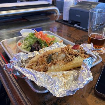 Backporch Drafthouse - Temple - A metal tray holds a taco wrapped in aluminum foil, accompanied by a small side salad with lettuce, tomato, and carrots, and a small container of dressing. A tall glass of dark soda with bubbles is placed nearby. The tray is on a wooden bar counter with bar equipment and a smartphone partially visible in the foreground. - American Near Me - Temple, Texas