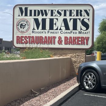 Midwestern Meats - A butcher shop that serves food - The sign for Midwestern Meats is mounted on a stone base and positioned outside a business establishment. It displays the business name in large, bold letters, with additional text indicating it is a restaurant and bakery. The sign also includes a logo with a pig's head and the phrase "Roger's Finest CornFed Meat!" The surrounding area shows a parking lot with a car partially visible, a beige wall, and some landscaping with plants and gravel. - Meat Shops Near Me - Mesa, Arizona