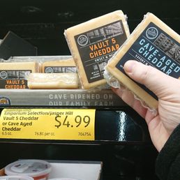 Aldis-Two different aged cheddar styles from Jasper Hill! (jaw drop) - A person is holding two blocks of Vault 5 Cheddar cheese from Jasper Hill in front of an ALDI grocery store shelf. The cheese blocks are labeled with black and orange or blue and white packaging. The shelf below displays a yellow price tag indicating $4.99 for the cheese, with other grocery items such as containers of salsa visible nearby.