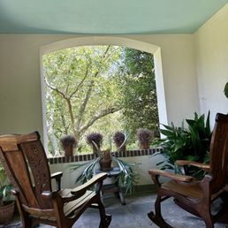 CHANTICLEER GARDEN - A porch area features two wooden rocking chairs positioned on a stone floor, with potted plants on either side. A large, arched window provides a view of trees and greenery outside, allowing natural light to fill the space. The ceiling is painted a light blue color, and the walls are white, creating a calm and inviting atmosphere.