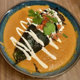 Chile Relleno - A ceramic bowl holds a serving of roasted eggplant covered in a smooth, orange-colored sauce. The dish is garnished with fresh cilantro leaves, diced tomatoes, and a drizzle of white cream sauce. The presentation emphasizes Mexican culinary elements, with the ingredients arranged on a wooden table.