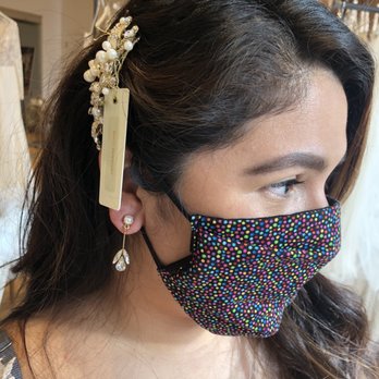 Jenny Yoo - Accessory try ons! - A woman with dark hair and earrings is wearing a multicolored polka dot face mask inside a retail store, with clothing racks in the background. - Bridal Near Me - Chicago, Illinois