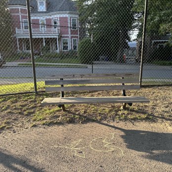 Schick Park - A wooden park bench with metal supports is placed on a dirt and grass patch in front of a chain-link fence. Behind the fence, residential houses with porches and trees are visible, indicating a neighborhood setting. The scene is illuminated by sunlight, casting shadows on the ground. - Playgrounds Near Me - Brookline, Massachusetts