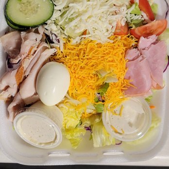 Davidos - Salad! - A takeout salad in a foam container contains sliced cucumbers, shredded cheese, chopped lettuce, tomato slices, ham, cooked chicken, a boiled egg, and two small containers of dressing. The ingredients are arranged separately within the container, which is open and viewed from above. The salad features a variety of fresh vegetables and protein options, suitable for a meal or snack. - Pizza Near Me - Rio Rancho, New Mexico