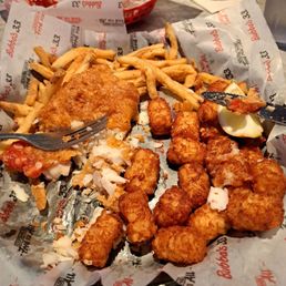 Fish and Chips - A basket lined with branded paper holds fried fish fillet, chicken nuggets, and French fries, with a lemon wedge on the side. The basket is on a table with a fork and a drink in the background, indicating a casual dining setting at Bubba's 33.