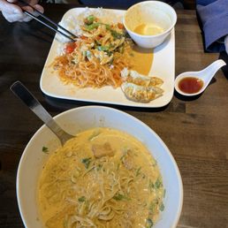 Pad Thai - The image shows two Asian noodle dishes on a dark wooden table. The larger bowl contains a creamy, yellowish noodle soup with visible green onions and pieces of meat. The second dish, served on a square plate, includes stir-fried noodles with vegetables, rice, and two gyoza dumplings, along with a small bowl of yellow curry or sauce and a small spoon of soy sauce. A person wearing a dark blue shirt is holding chopsticks over the second dish.