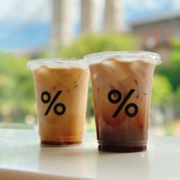 Kyoto Latte - Two iced coffee beverages in clear plastic cups with a black percentage logo are positioned on a white surface, with a blurred outdoor background.