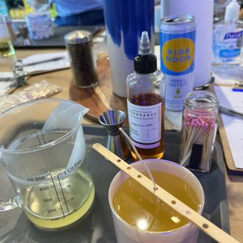 Candles Off Main - A table at a candle-making workshop features various supplies such as a measuring cup with yellow liquid, a container with a wick, fragrance oil, matches, and other tools. The background shows additional workshop items, a person in a blue shirt, and a roll of paper towels. The setup indicates an interactive candle-making activity. - Candle Stores Near Me - Annapolis, Maryland