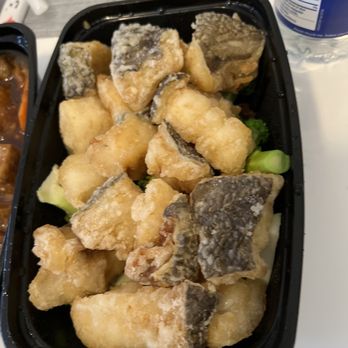 Congee Queen - Cod - The image shows a black plastic takeout container filled with battered and fried fish pieces. The fish has a golden, crispy coating, and some pieces have visible fish skin. There is a small amount of green garnish, possibly lettuce, underneath the fried fish. A bottle of water and part of another dish are visible on the table. - Chinese Near Me - Markham, Ontario
