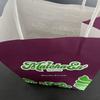 Matcha En - A paper shopping bag from Matcha En features green and purple branding, with the website URL and Japanese characters, placed on a dark surface. - Desserts Near Me - Champaign, Illinois