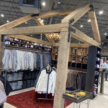 Boot Barn - The image shows a clothing display inside a retail store, featuring racks of shirts, jackets, and hats. The display is framed by a rustic wooden structure with visible metal brackets. A small table with tools is positioned nearby, and a red rug is on the floor beneath the display. In the background, other clothing racks and customers are visible, indicating a large retail environment with ample lighting and organized shelving. - Shoe Stores Near Me - Bridgeville, Pennsylvania