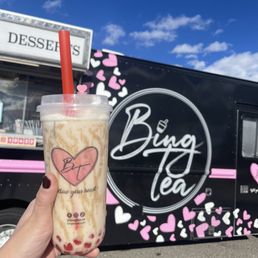 Gingerbread milk tea with boba - A person holds a bubble tea with a red straw in front of a Bing Tea food truck decorated with pink and white hearts, under a blue sky.