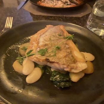 Cafe Vincotto - Branzino - A serving of branzino fish is presented on a black plate, accompanied by white beans and leafy greens. The fish is garnished with microgreens, and the dish appears to be part of a fine dining experience, with a glass of water and other plates visible in the background. - Desserts Near Me - Berkeley Heights, New Jersey