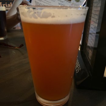 Murphys Irish Pub - Firestone Walker Cali Squeeze Blood Orange Hefeweizen - A pint glass containing Blood Orange Hefeweizen beer is placed on a dark table at Murphys Irish Pub, with menus and a person in the background. - Irish Pub Near Me - Murphys, California