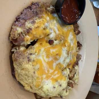 Eli's - A breakfast dish featuring a large omelette topped with melted cheddar cheese and pieces of bacon. The omelette appears fluffy and cooked thoroughly. A small black cup of ketchup is placed on the side of the beige plate. The plate is on a light-colored table surface, with some crumbs and food remnants visible around the dish. - Breakfast & Brunch Near Me - Natick, Massachusetts