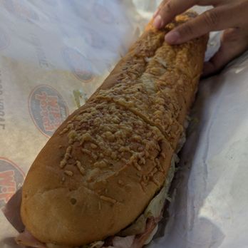 Jersey Mike's Subs - A long sub sandwich with melted cheese, meats, and lettuce on a toasted bun, partially wrapped in branded paper, being held by a person's hand. - Sandwiches Near Me - Onalaska, Wisconsin