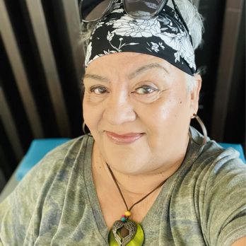 Macy's Backstage - See how beautiful I look with my facial makeover. I am loving it Have a wonderful blessed day - A woman with gray hair, partially covered by a black and white floral headscarf, is taking a selfie. Sunglasses rest on her head, and she is wearing a gray T-shirt. She has a large, round green pendant necklace with a decorative element and small colorful beads. The background includes black vertical bars and a blue surface, suggesting an indoor setting. - Women's Clothing Near Me - Montebello, California