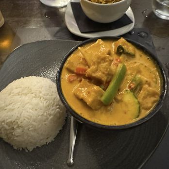 Red Torch Ginger - Red Thai curry - The image shows a meal with a serving of white rice, a bowl of curry containing vegetables and tofu in a yellow sauce, and a bowl of noodles with garnishes in the background. The dishes are placed on a dark plate and table, with glasses of water nearby. - Thai Near Me - Dublin, D