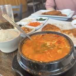 Soondubu, 5 banchan of the day for $5 - The image shows a traditional Korean meal with a hot stone bowl of spicy soup, garnished with chopped green onions. A bowl of steamed white rice is placed nearby. In the background, there are multiple small dishes containing side items such as vegetables and pickles, along with other plates of food. The setting appears to be a casual Korean restaurant with wooden decor and tables.