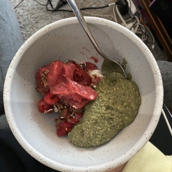 The Dripping Root - Double Date smoothie bowl and matcha chia seed pudding - A ceramic bowl contains a smoothie bowl topped with red fruit pieces and a green chia seed pudding. A metal spoon is inserted into the bowl. The background includes a gray carpet, electronic cords, and a wooden shelf with items. The person holding the bowl is wearing a yellow sleeve, and the setting appears to be indoors, possibly in a home or cafe environment. - Juice Bars & Smoothies Near Me - Minneapolis, Minnesota