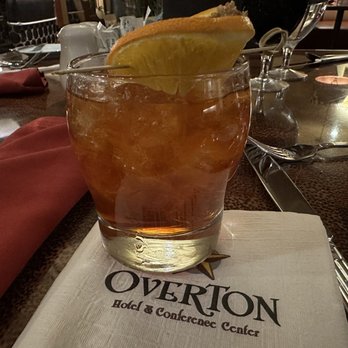 Pecan Grill - Maple Pecan Old Fashioned (12/23/2022) - A glass of amber-colored cocktail with ice and a lemon wedge garnish is placed on a napkin bearing the Overton Hotel & Conference Center logo. The table setting includes utensils and a reflective surface, indicating a dining environment at the hotel restaurant. The background shows a dimly lit, upscale dining area with additional glassware and tableware. - New American Near Me - Lubbock, Texas