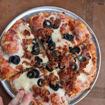 Roaring Rapids Pizza Co - Kids pizza (chubby toddler hand for scale) - A pizza with a cheese base, topped with bacon bits and sliced black olives, cut into four slices and placed on a round metal tray. A child's hand is visible at the bottom left corner of the image, touching the tray. - Pizza Near Me - Eugene, Oregon