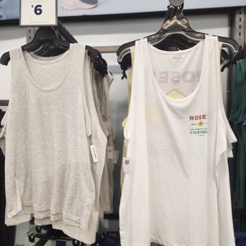 Old Navy - The image shows a retail clothing display with sleeveless tank tops hanging on black hangers. One tank top is light gray, and another is white with printed text and graphics. A price sign above indicates the current price as $6. The background includes a blurred image of a person walking, suggesting the store environment. The display is organized with additional clothing items visible below. - Men's Clothing Near Me - Davie, Florida