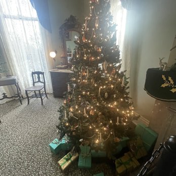 David Davis  Mansion - A Christmas tree decorated with string lights, ornaments, and candles stands in a vintage-style room. The room features lace curtains, an antique mirror, and classic furniture, including a small table and chairs. Several wrapped presents are placed under the tree, and sunlight filters through the lace curtains, creating a cozy holiday scene. - Museums Near Me - Bloomington, Illinois