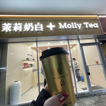 Molly Tea - A person with painted nails holds a coffee cup in front of Molly Tea, a tea shop with illuminated signage displaying Chinese characters and the name 'Molly Tea.' The shop has a glass storefront with visible interior lighting and a few customers inside. The scene is set outdoors on a paved sidewalk at night. - Bubble Tea Near Me - Markham, Ontario