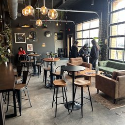 A coffee shop interior features various tables and chairs, pendant lights, and large windows with natural light, creating a cozy atmosphere.