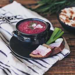 Borsch - A black bowl filled with Ukrainian borscht, a beet-based soup, topped with sour cream and garnished with green onions. The bowl is placed on a black plate, accompanied by slices of bread with bacon and a whole green onion. The setting includes a striped cloth on a wooden surface, with additional food items visible in the background.