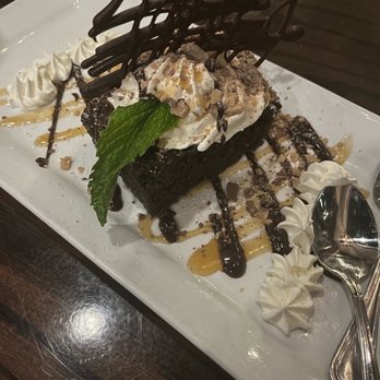 Buck & Honey's - Better than Chocolate Cake - A chocolate brownie dessert topped with whipped cream, a mint leaf, and chocolate drizzle, served on a white rectangular plate with decorative whipped cream. - American Near Me - Monona, Wisconsin