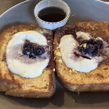 The Brunch Pub - Lemon Ricotta French Toast - Two slices of French toast are served on a plate, each topped with whipped cream, blueberries, and lemon zest. A small bowl of syrup is placed above the toast. The dish appears to be part of a breakfast or brunch meal. - Pubs Near Me - Centerville, Ohio