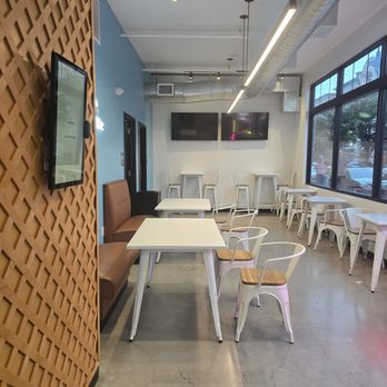 Wondaffle - The image shows a cafe interior with a mix of seating options, including white tables with metal chairs that have wooden seats, and a brown upholstered bench along a blue accent wall. Two large flat-screen monitors are mounted on the wall at the back. The space has high ceilings with exposed ductwork and a long, linear ceiling light. Large windows on the right side provide natural light and a view of parked cars and trees outside. The floor is polished concrete, and the overall decor is modern and minimalistic. - Waffles Near Me - Bethlehem, Pennsylvania