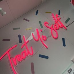 A neon sign with the phrase 'Treat Yo Self' in pink is mounted on a wall. The wall is decorated with various pastel-colored sprinkle shapes, creating a playful and inviting atmosphere. The sign is illuminated, making it stand out against the background.