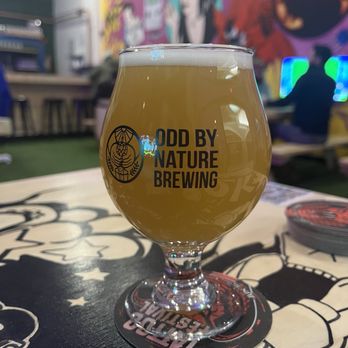 Odd By Worcester Brewing - Nature... - A beer glass with a hazy, yellowish beer, featuring the Odd By Nature Brewing logo, is placed on a table inside a brewery with colorful murals in the background. - Beer Gardens Near Me - Worcester, Massachusetts