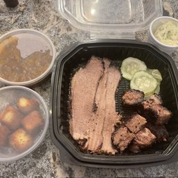 Burnt Ends Sandwich - A barbecue meal includes sliced brisket, burnt ends, cucumber slices, coleslaw, and fried potatoes in plastic containers on a granite countertop.