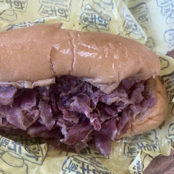 The Hat - Pastrami Dip. - A sandwich featuring shredded meat, possibly roast beef or brisket, inside a soft, slightly shiny bun. The sandwich is resting on yellow paper with printed logos and text, indicating it is from a fast-food establishment. The bun appears fresh, and the meat is piled generously, with some juices visible. - American Near Me - Pasadena, California