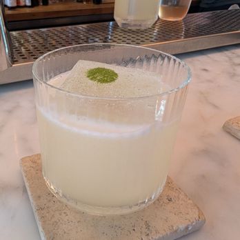 Bar Pompette - Their version of a margarita, it was wonderful - A pale-colored cocktail with a frothy surface and a green powder garnish is served in a clear glass on a marble table at a bar. - Cocktail Bars Near Me - Toronto, Ontario