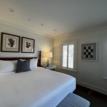 Harbor View Hotel - Room - The image shows a bedroom with white paneled walls and a ceiling with recessed lighting. A bed with white linens and a striped pillow is positioned against the wall, with a dark wooden headboard. Next to the bed is a white nightstand with a lamp that has a white shade. There are three framed botanical prints above the bed and a framed geometric artwork on the wall near a window with white shutters. The room has a dark wood floor and a bench at the foot of the bed with a patterned fabric cover. - Hotels Near Me - Edgartown, Massachusetts